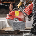 A red and black Hitachi stone saw in use on a wooden pallet, cutting a large gray granite block at a construction site.