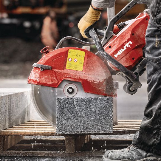 A red and black Hitachi stone saw in use on a wooden pallet, cutting a large gray granite block at a construction site.